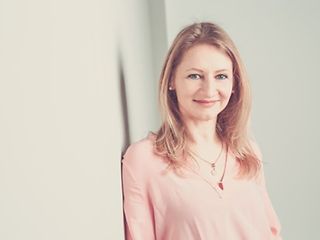 Eine junge Frau lehnt lächelnd an einer hellen Wand und blickt in die Kamera.