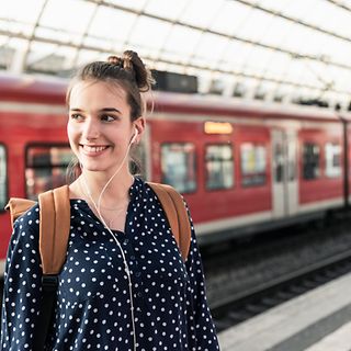 Eine junge Frau mit Rucksack und Kopfhörern steht am Gleis eines Bahnhofs.