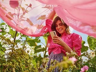 A woman with a pink vail is standing in a flower field. She is holding a smartphone.