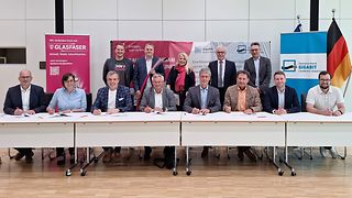 Vertreter der Kommunen sitzen mit Unterschriftenmappen an einer Tischreihe, die übrigen Personen stehen dahinter.