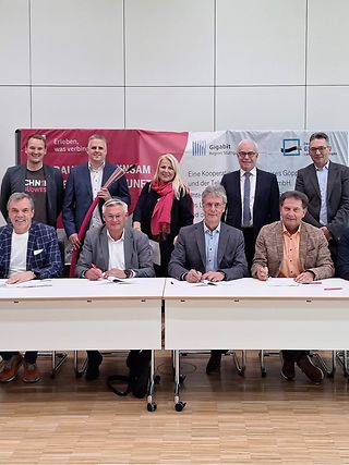 Vertreter der Kommunen sitzen mit Unterschriftenmappen an einer Tischreihe, die übrigen Personen stehen dahinter.